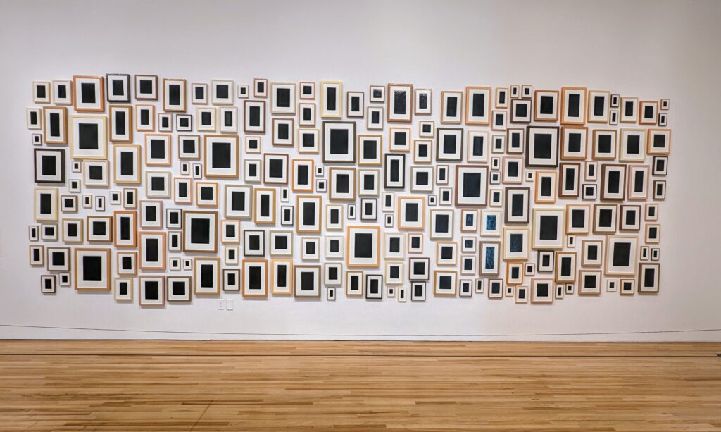 Art installation by Allan McCollum. Dozens of painted black canvases of similar but different shapes and sizes. All with similar but different frames.